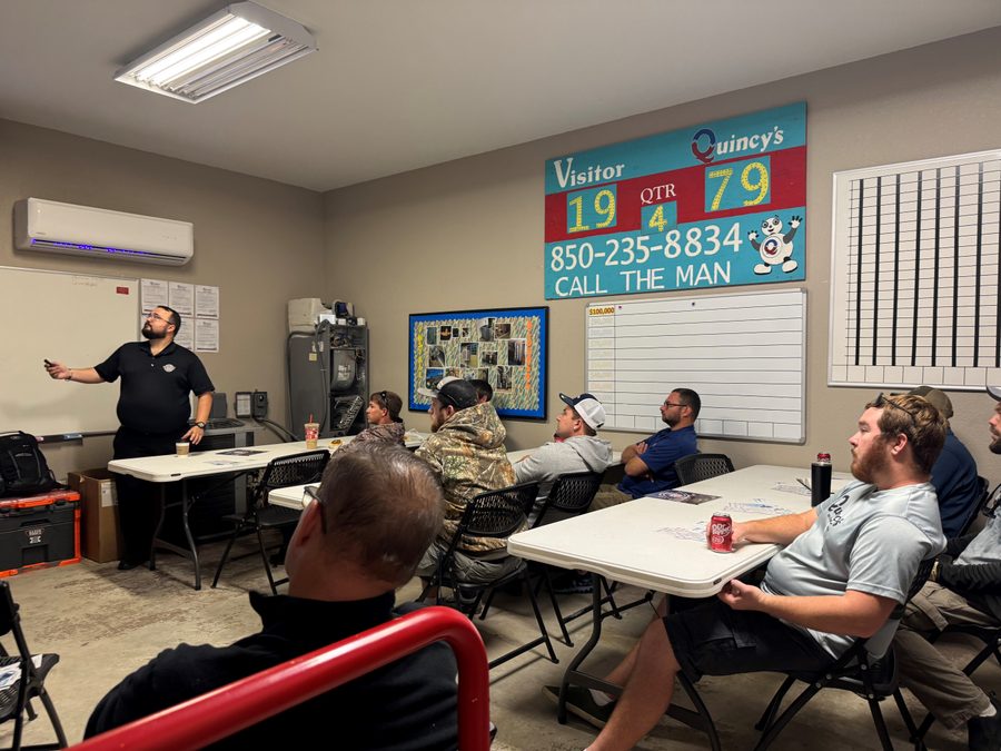 Quincy's team at an HVAC training session — scoreboard on the wall reads 'Quincy's 79, Call the Man, 850-235-8834'